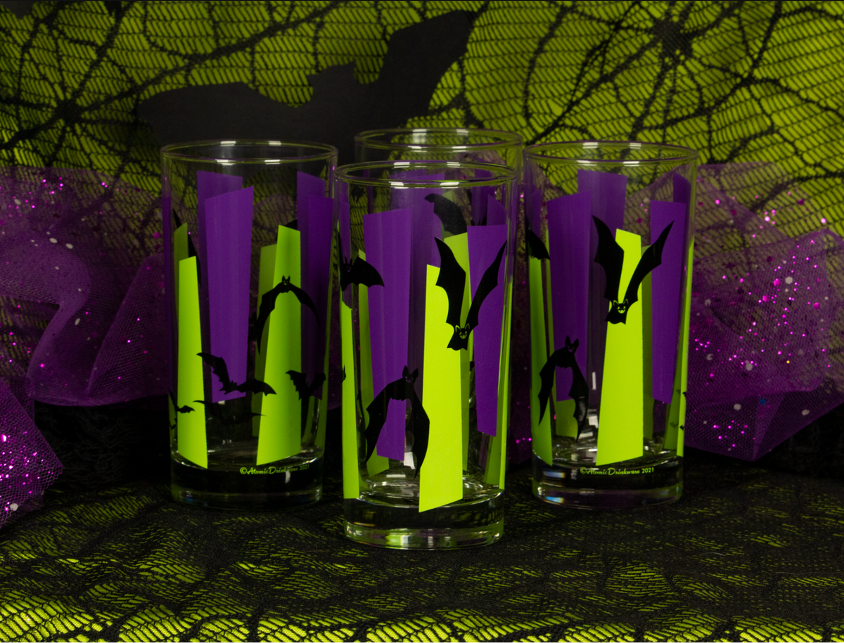 Clear glass tumblers printed with purple and chartreuse green vertical googies and black bat silhouettes, set against a Halloween-themed background of black lace, purple mesh, and a green spiderweb pattern.