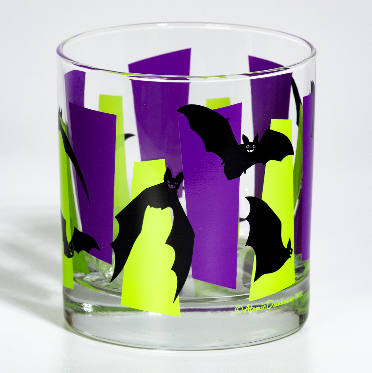 A clear glass tumbler with a Halloween design featuring black bats flying over purple and neon green geometric googies, photographed on a white background.