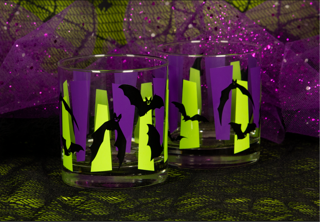 Two clear glass tumblers with Halloween-themed bat silhouettes on purple and chartreuse green retro googies. set against a spiderweb fabric and purple tulle backdrop.