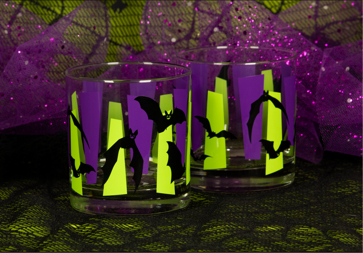 Two clear glass tumblers with Halloween-themed bat silhouettes on purple and chartreuse green retro googies. set against a spiderweb fabric and purple tulle backdrop.