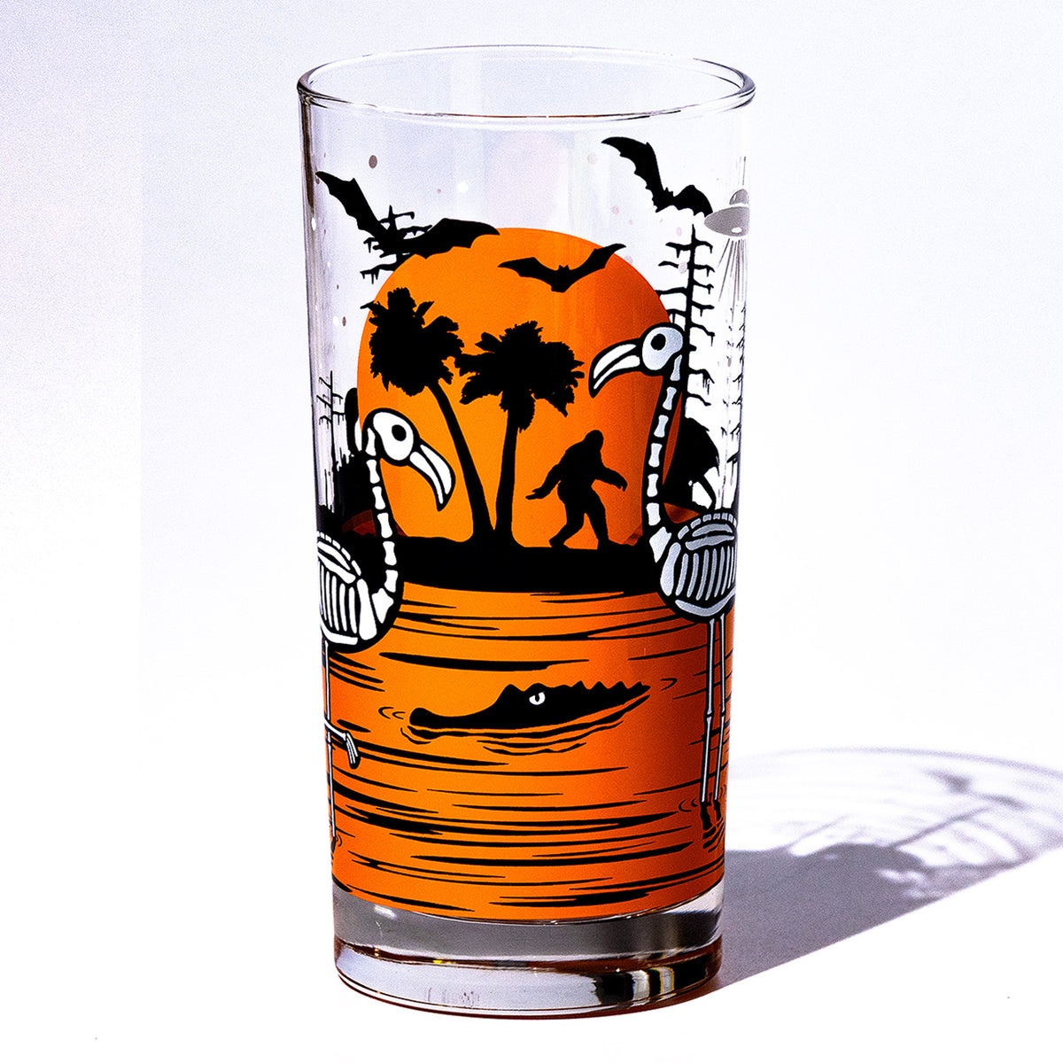 Orange Eerie Everglades Collins single glass with black spooky swamp silhouettes and glow-in-the-dark skeleton flamingos, skelemingos, on a white background.