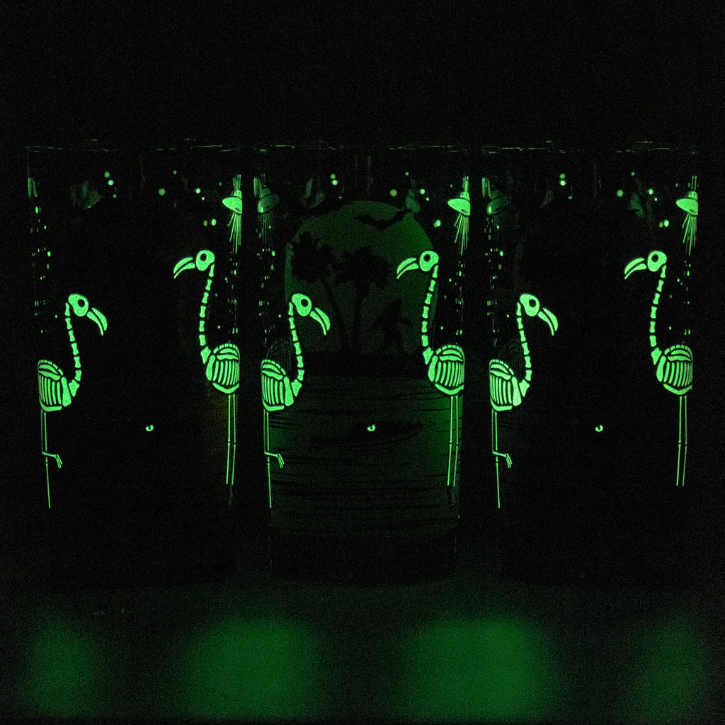Glow-in-the-dark glass cups decorated with glowing skeleton flamingo skelemingo designs, shown in a dark environment to highlight their luminous effect.