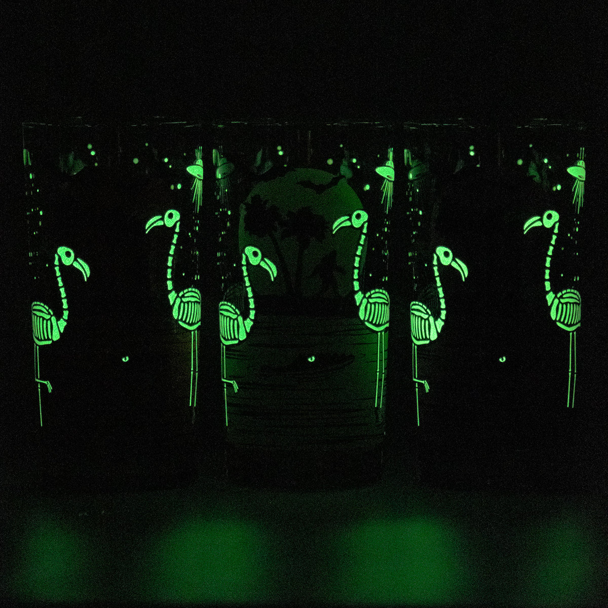 Glow-in-the-dark glass cups decorated with glowing skeleton flamingo skelemingo designs, shown in a dark environment to highlight their luminous effect.
