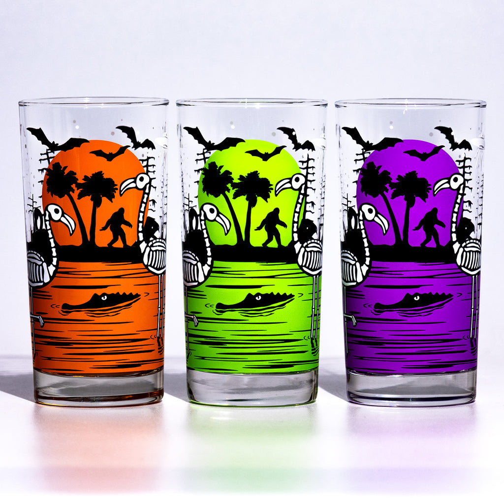 Line up of three Eerie Everglades Collins glasses. One orange, one chartreuse and one purple glass with black spooky swamp silhouettes and glow-in-the-dark skeleton flamingos, skelemingos. One a white background.