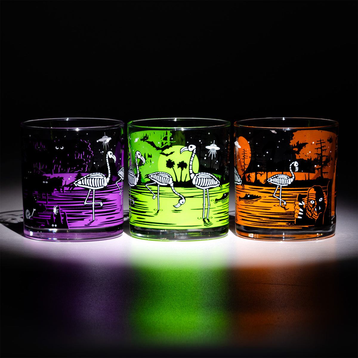 Three clear rocks glasses with colorful skeleton flamingo designs, shown lit up against a black background. The left glass glows purple, the middle glass glows green, and the right glass glows orange.