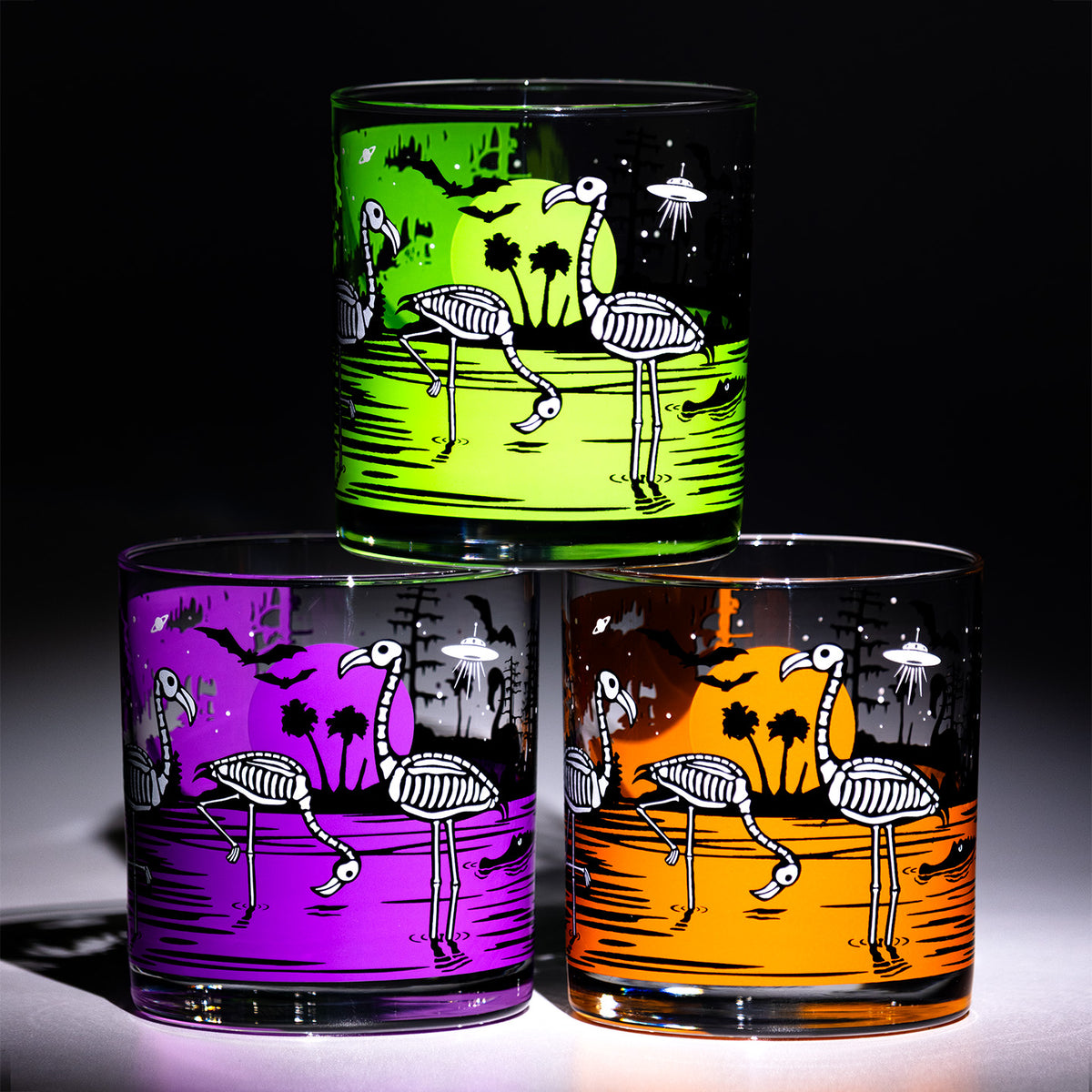Three clear rocks glasses with different vibrant color backgrounds (green, purple, and orange) featuring a glow in the dark skeleton flamingo and palm tree design, arranged in a pyramid against a black background.