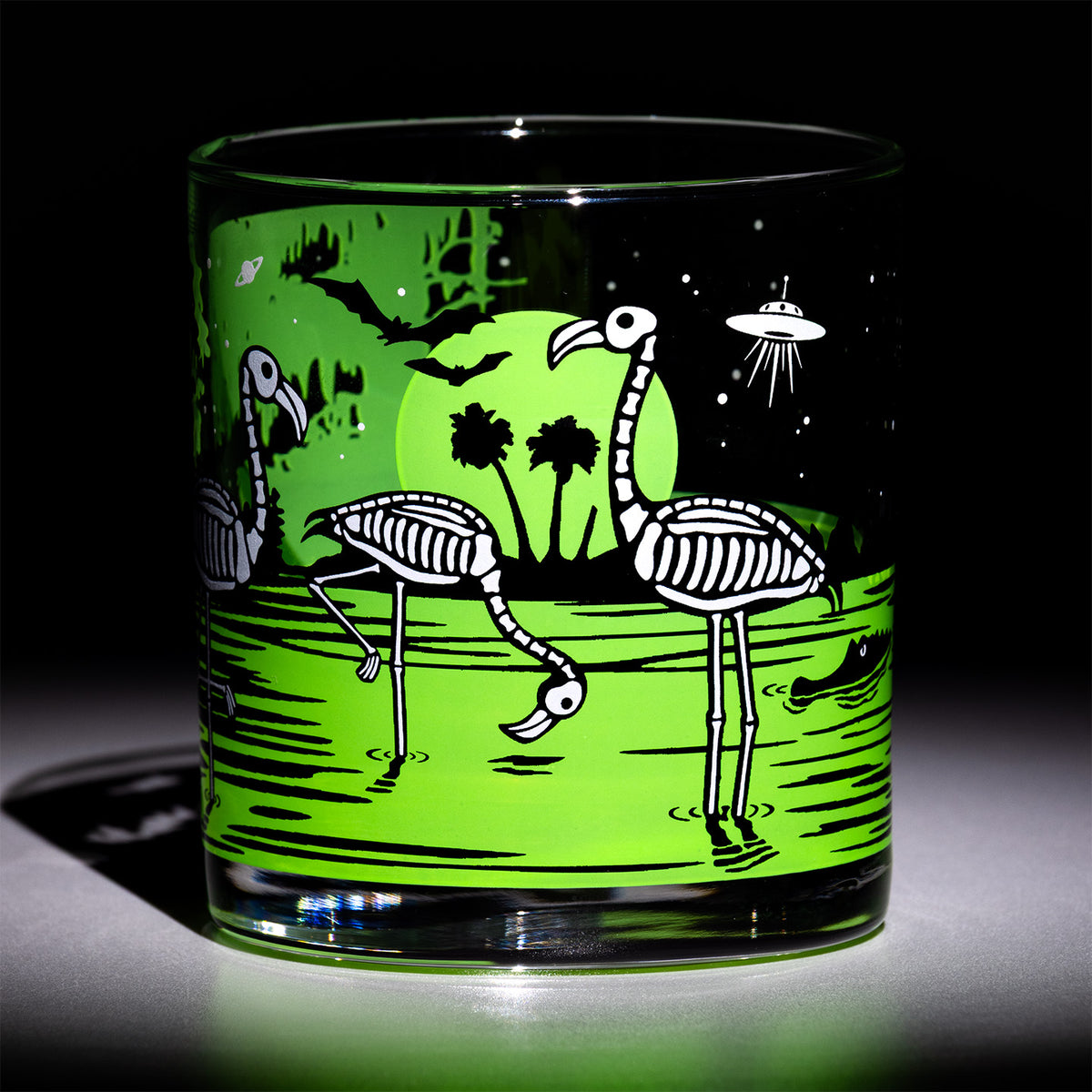 A clear glass tumbler with a printed graphic of green swamp, skeleton flamingos, black silhouettes, and a UFO against a bright neon green and black background, photographed on a dark studio background.