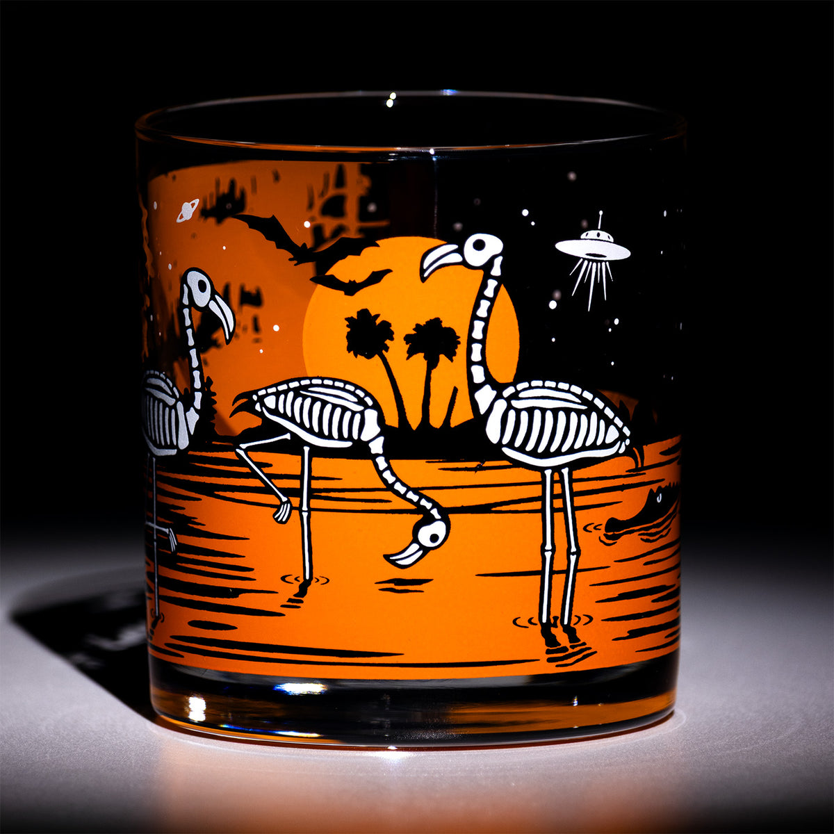 A clear glass tumbler with a printed Halloween-themed design featuring skeleton flamingos, palm trees, a black sky, and a flying saucer over an orange moon and water, photographed against a dark background.