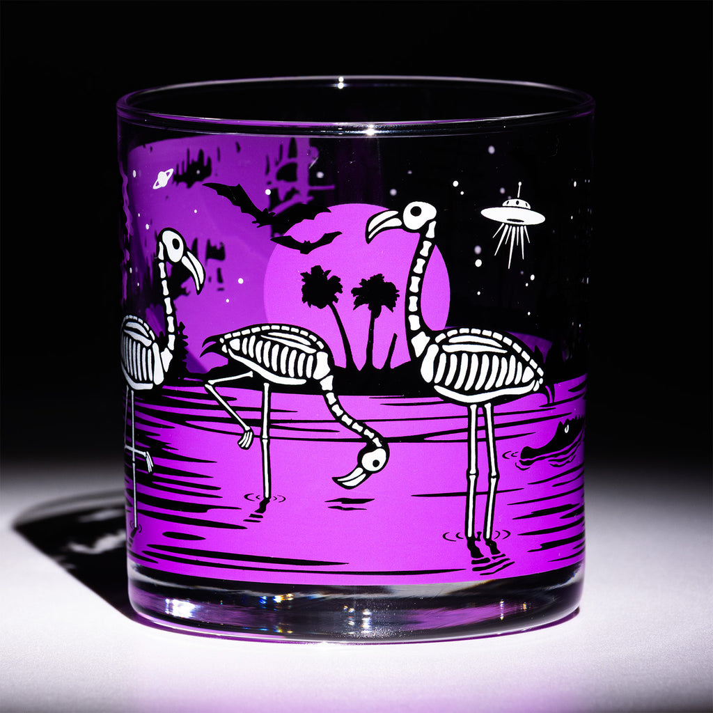 A clear glass rocks tumbler featuring a purple and black graphic print of skeleton flamingos, palm trees, a UFO, and bats on a dark background.