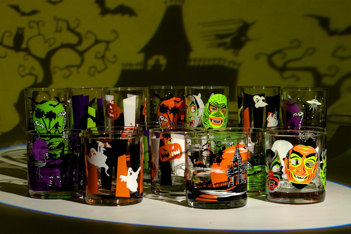 A set of Halloween-themed Collins and rocks glasses arranged on a white surface, featuring spooky illustrations like pumpkins, ghosts, witches, and a haunted house. The glasses are shown against a dark green background with black silhouettes of a haunted house, bats and a tree.