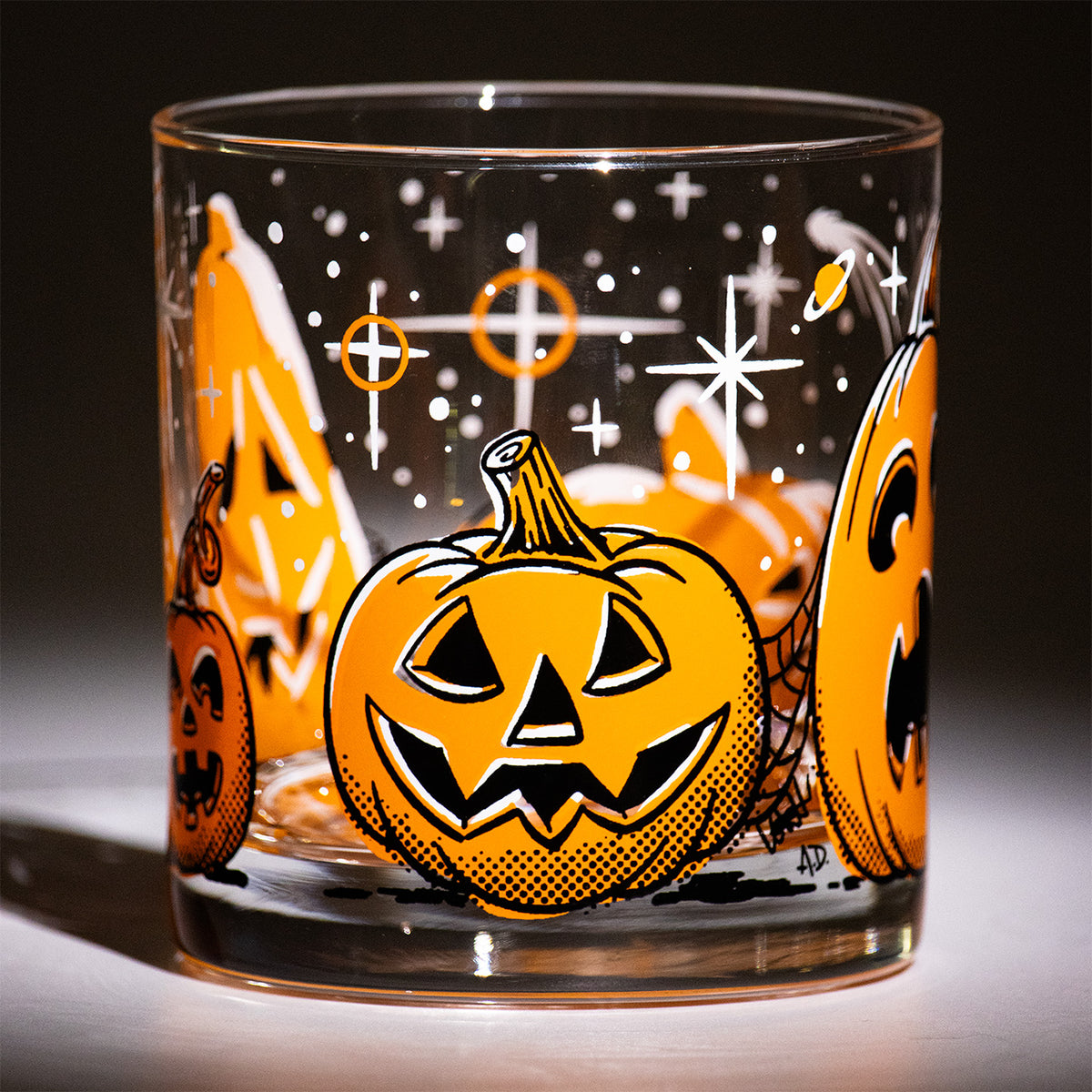 Five Little Pumpkins Rocks glass with retro style hand illustrated jack-o-lantern with shooting stars on a dark background.  Friendly round Halloween pumpkin with triangle eyes and nose