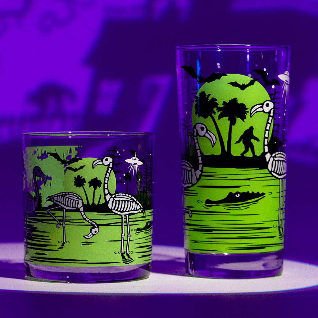 2 UV Fluorescent chartreuse green Eerie Everglades Collins and Rocks glasses with black spooky swamp silhouettes and glow-in-the-dark skeleton flamingos, skelemingos, on a purple spooky Halloween haunted house shadow background.