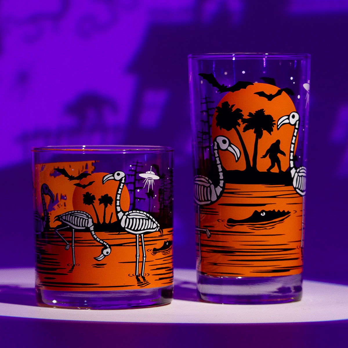 Two Halloween-themed drinking glasses with orange, black, and white illustrations of flamingos, bats, an alligator, swamp ape, and a silhouetted landscape, displayed on a white surface against a purple-lit background.
