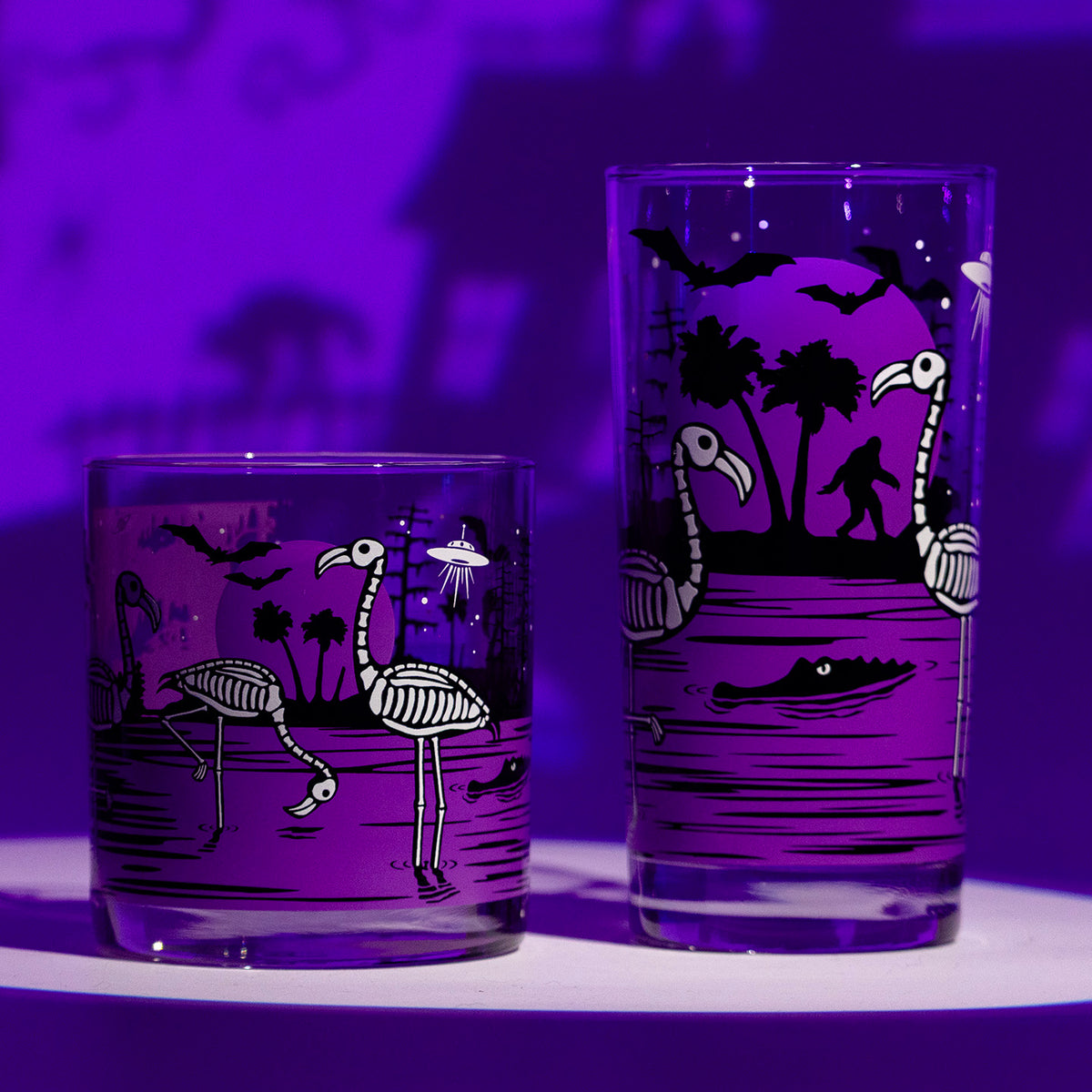 2 UV Fluorescent purple Eerie Everglades Collins and Rocks glasses with black spooky swamp silhouettes and glow-in-the-dark skeleton flamingos, skelemingos, on a purple spooky Halloween haunted house shadow background.