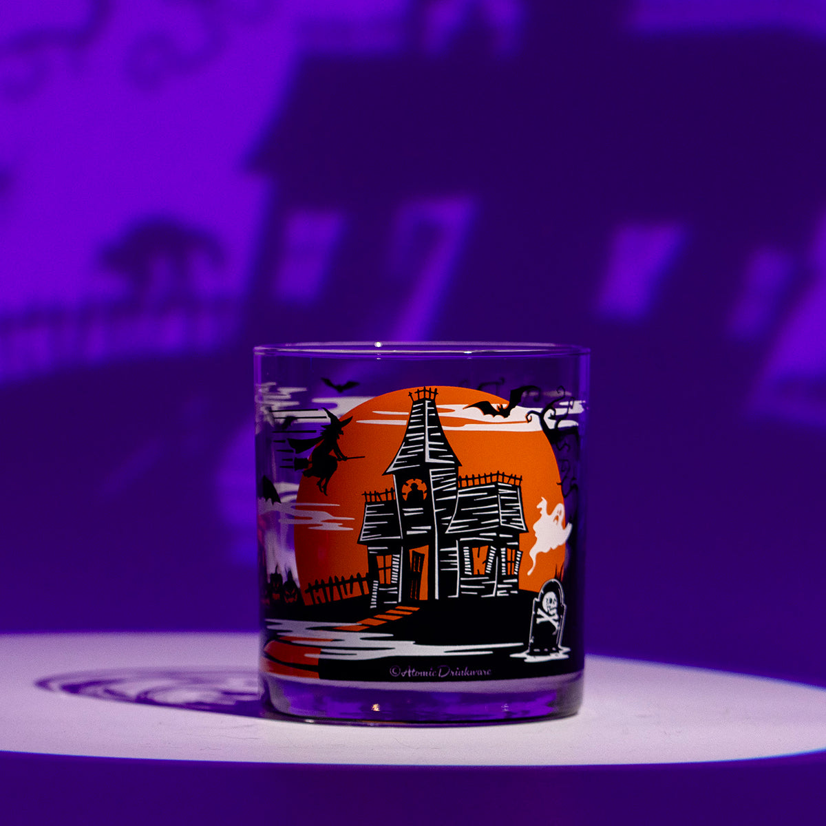 A clear glass rocks tumbler with a hand screen-printed Halloween design featuring a haunted house, bats, a large orange moon, ghost, witch, gravestone and black cat, photographed on a white surface against a purple-lit background.