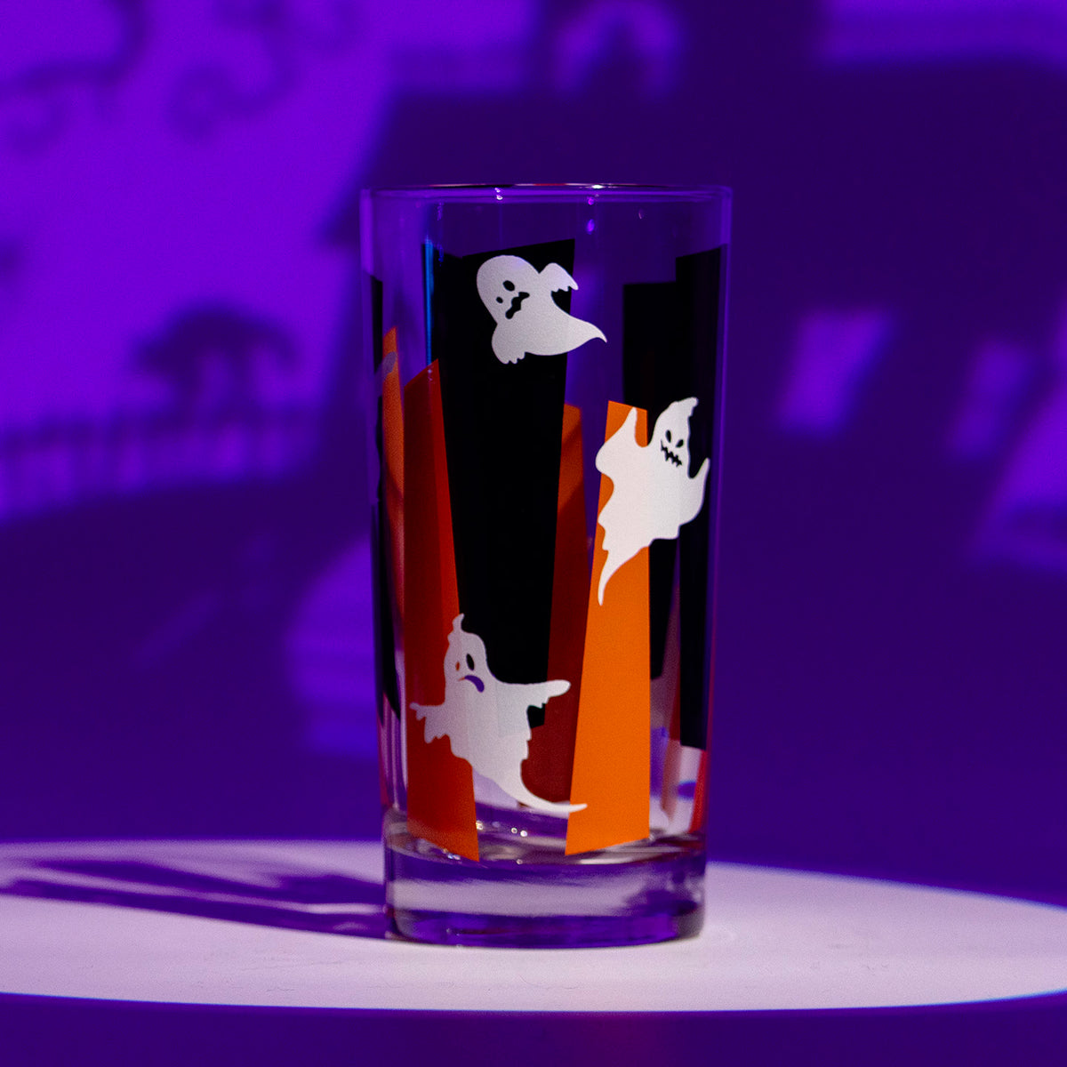 A clear drinking glass with Halloween-themed glow in the dark ghosts set against orange and black googie geometric shapes. Photographed with a purple background with dramatic lighting.