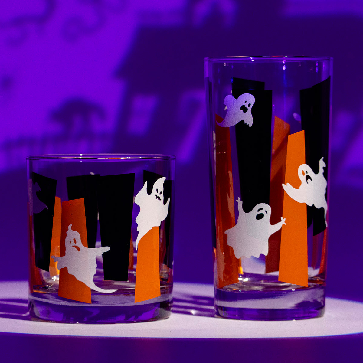 Two clear glass tumblers with Halloween-themed glow in the dark ghosts and orange and black googie designs displayed on a white surface under purple ambient lighting.