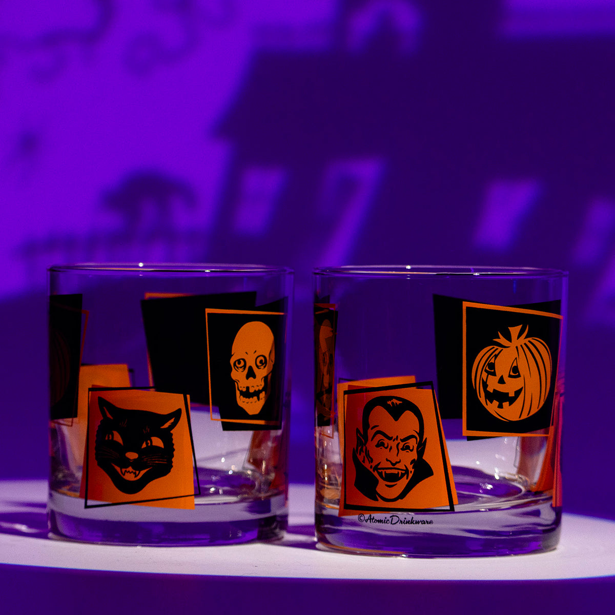 2 Monster Mash Rocks glasses with retro orange and black Halloween screen-printed designs, owl, bats, black cat, skull, jack-o-lantern, and vampire on a Halloween themed purple background.