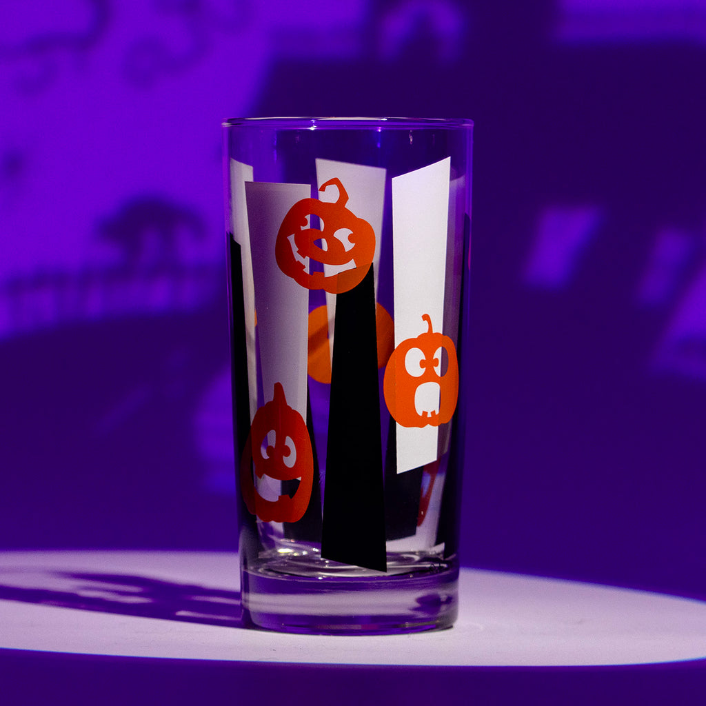 A clear drinking glass decorated with Halloween themed orange jack-o'-lantern pumpkins over black and white vertical googie stripes, set against a purple-lit background.