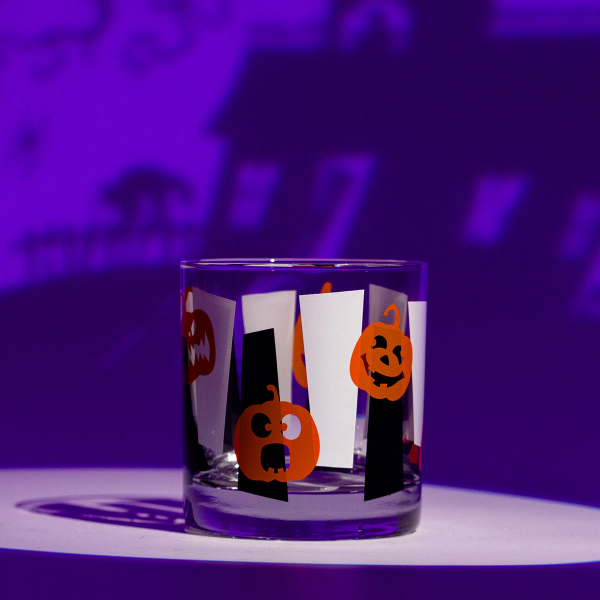 A clear glass tumbler with Halloween-themed orange jack-o'-lanterns pumpkins printed on black and white googies, set against a purple-lit background with a house silhouette.