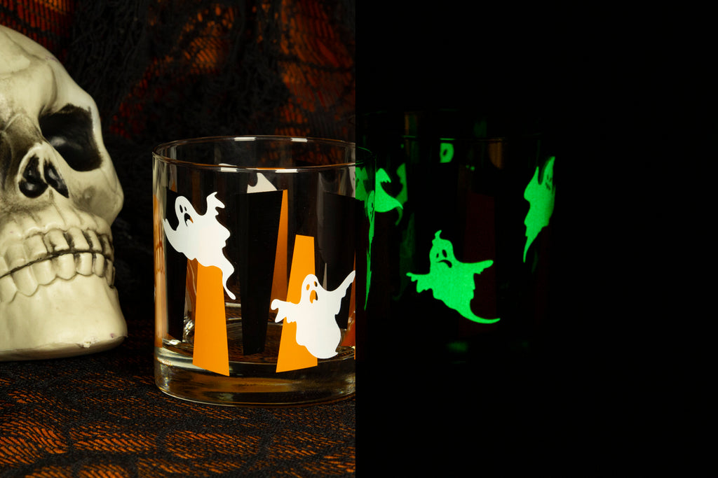 A clear glass tumbler with Halloween ghost and orange and black googie stripes, shown beside a skull prop on a spiderweb tablecloth, with a black-and-dark scene on the right showing the ghosts glowing green in the dark.