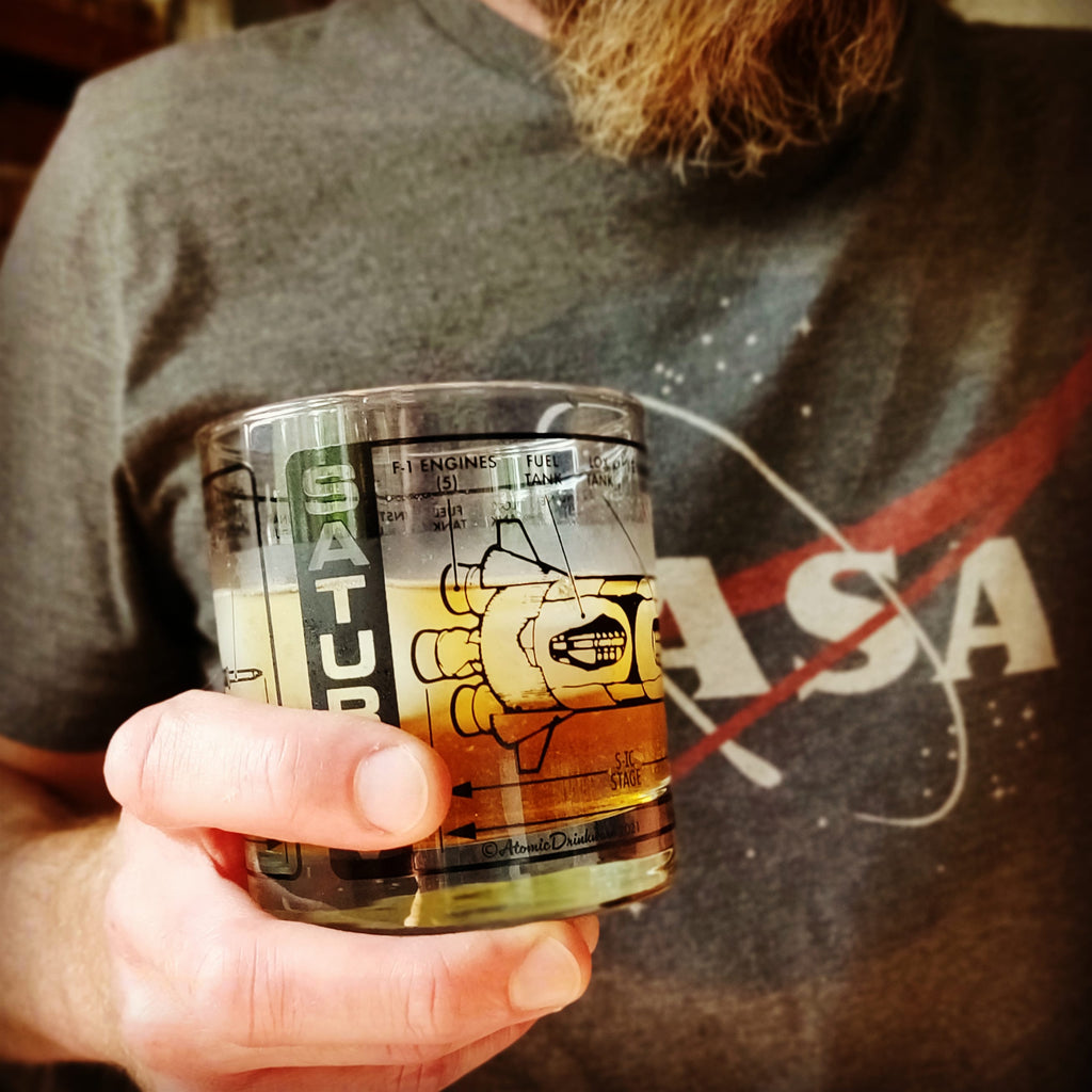 Person holding a glass with a NASA-themed retro cocktail glass that says Saturn V, wearing a NASA shirt.