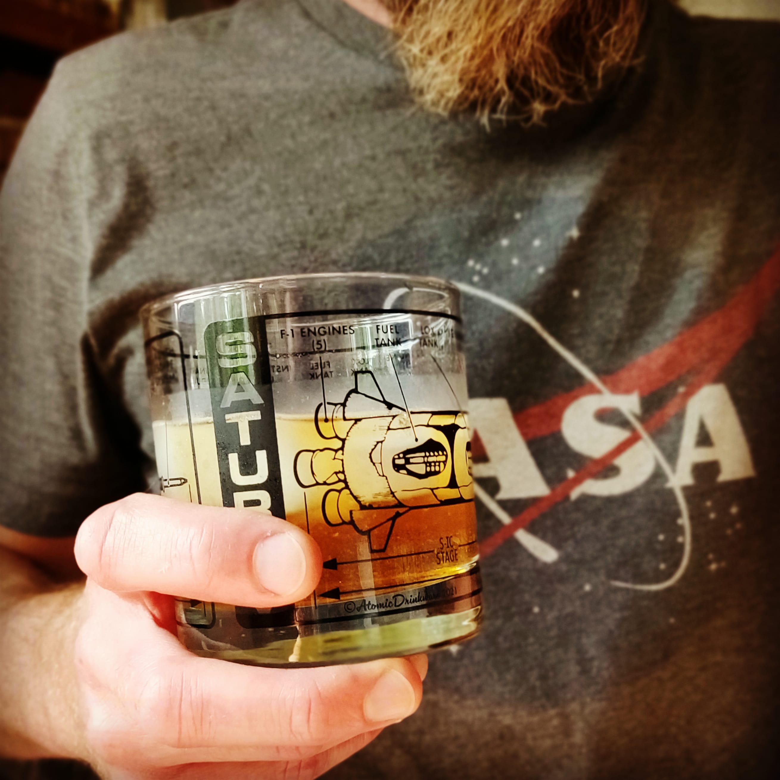 Person holding a glass with a NASA-themed retro cocktail glass that says Saturn V, wearing a NASA shirt.