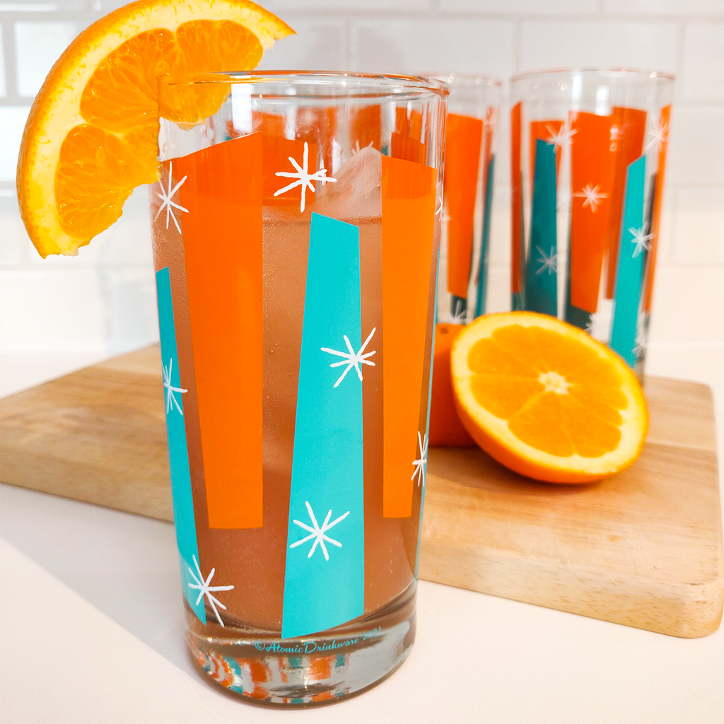 Glass with a colorful mid century design, filled with a beverage and an orange slice, on a wooden cutting board.
