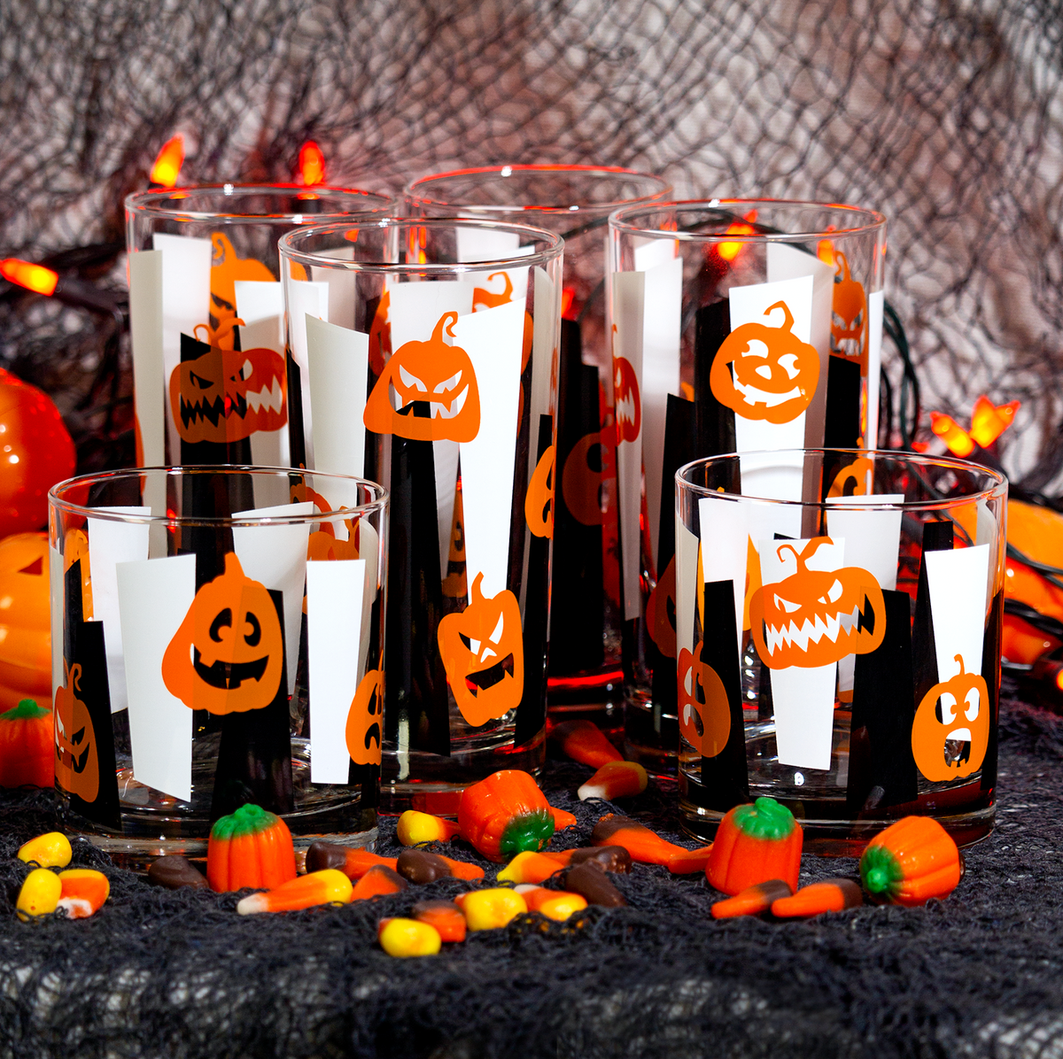 Collins and Rocks drinking glasses printed with orange jack-o'-lantern faces over black and white vertical googie stripes, arranged on a black webbed surface with Halloween decorations, candy corn, and orange pumpkins in the background.