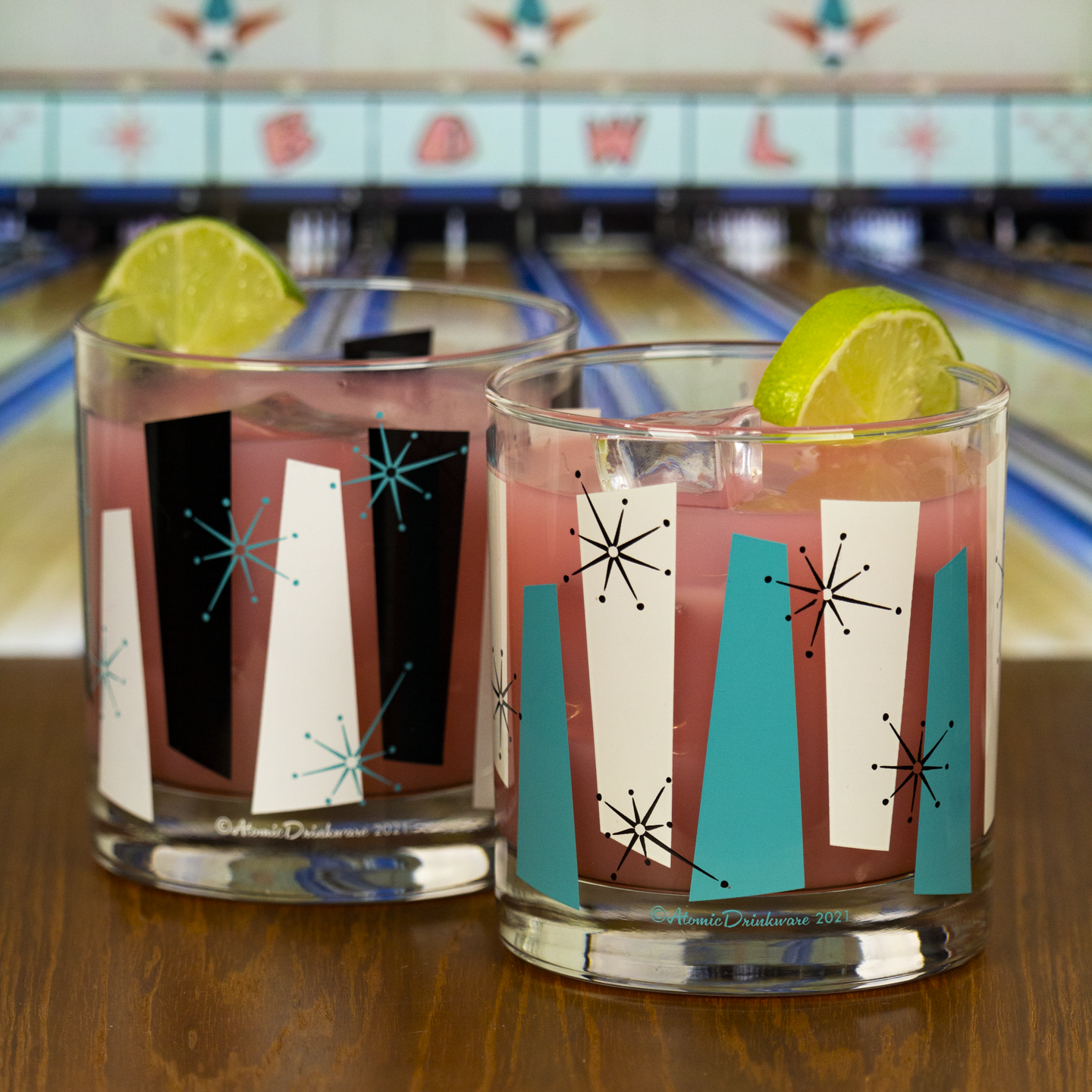 Two retro-style glasses with colorful rock a billy designs on a bowling alley counter.