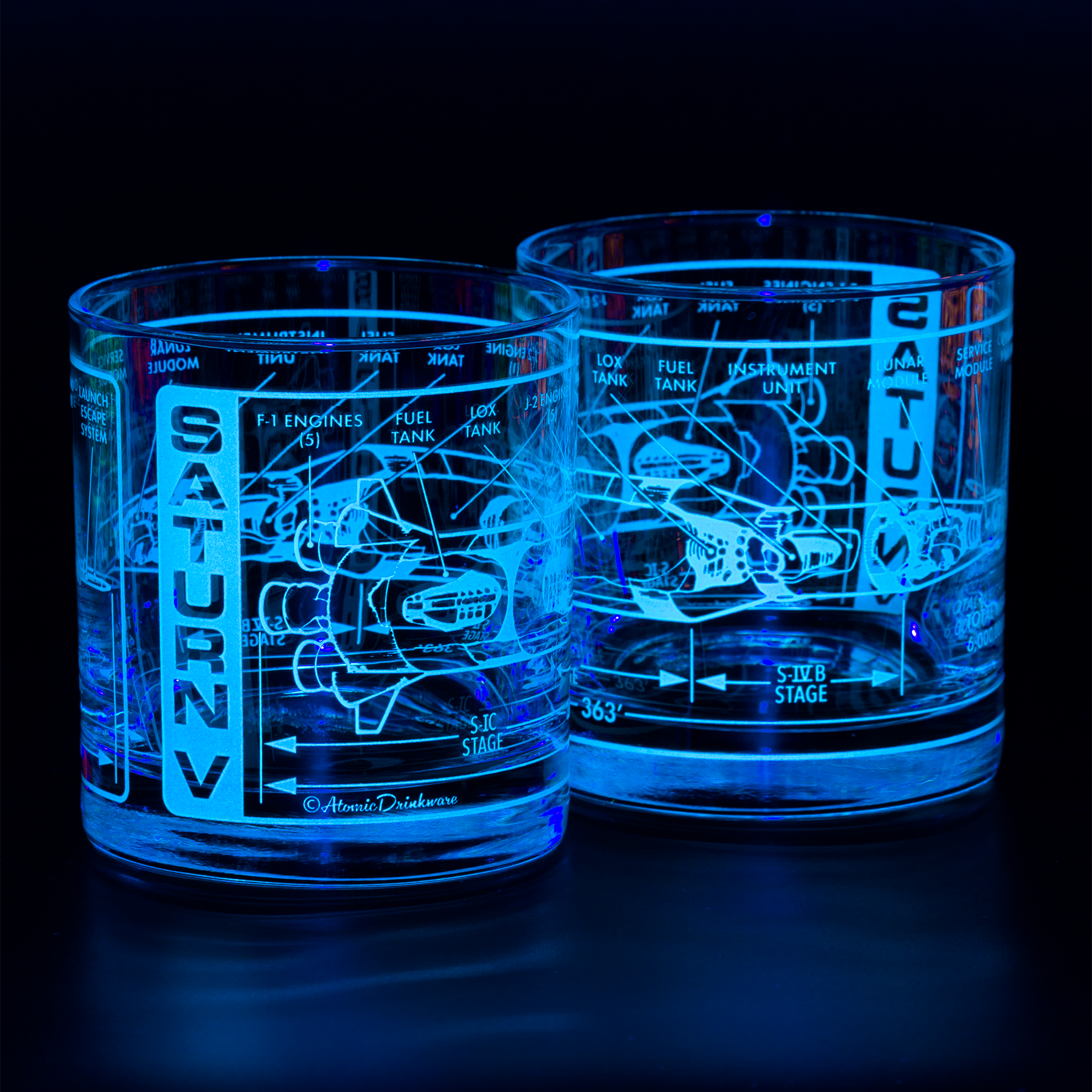 Two illuminated glass tumblers with Saturn V rocket blueprint engraving on a dark background