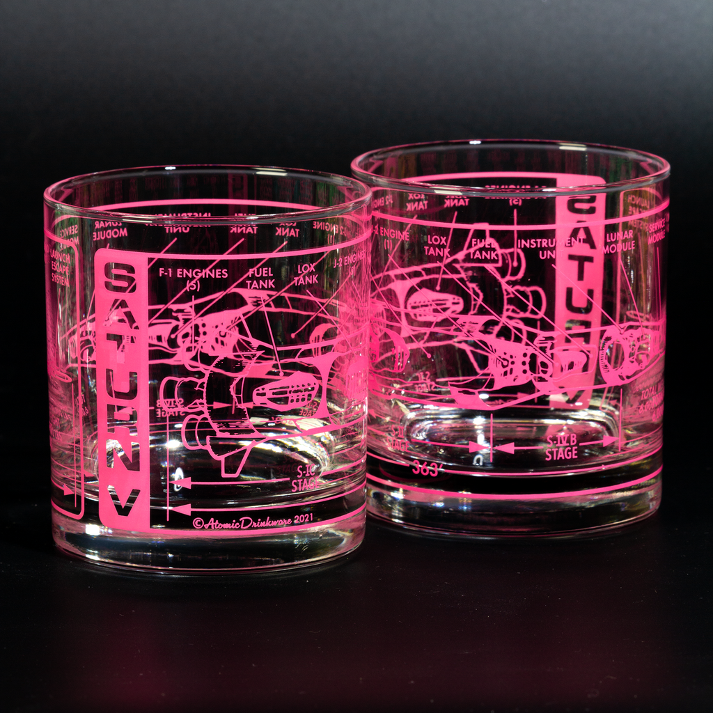 Two glass tumblers with engraved space technical drawings illuminated by a pink light on a black background.