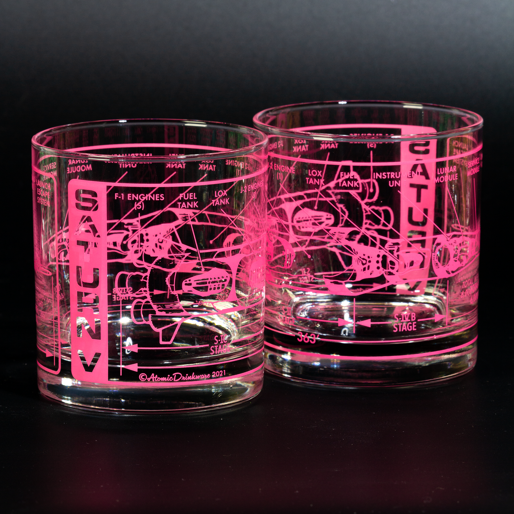 Two glass tumblers with engraved space technical drawings illuminated by a pink light on a black background.