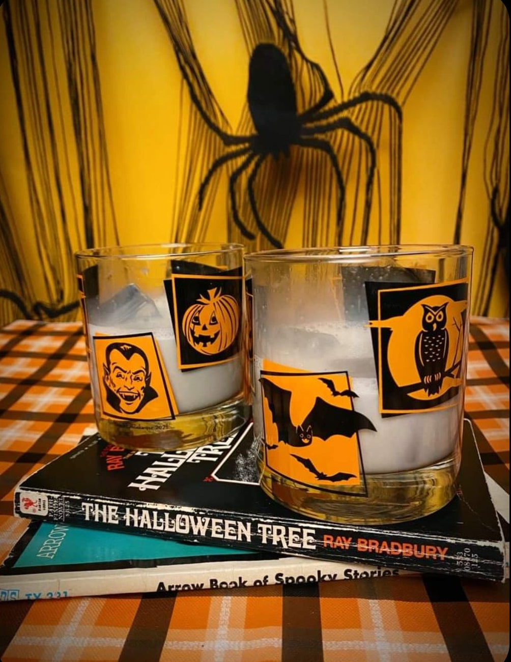 2 Monster Mash Rocks glasses with retro orange and black Halloween designs, owl, bats, jack-o-lantern, and vampire on a Halloween themed table with books.