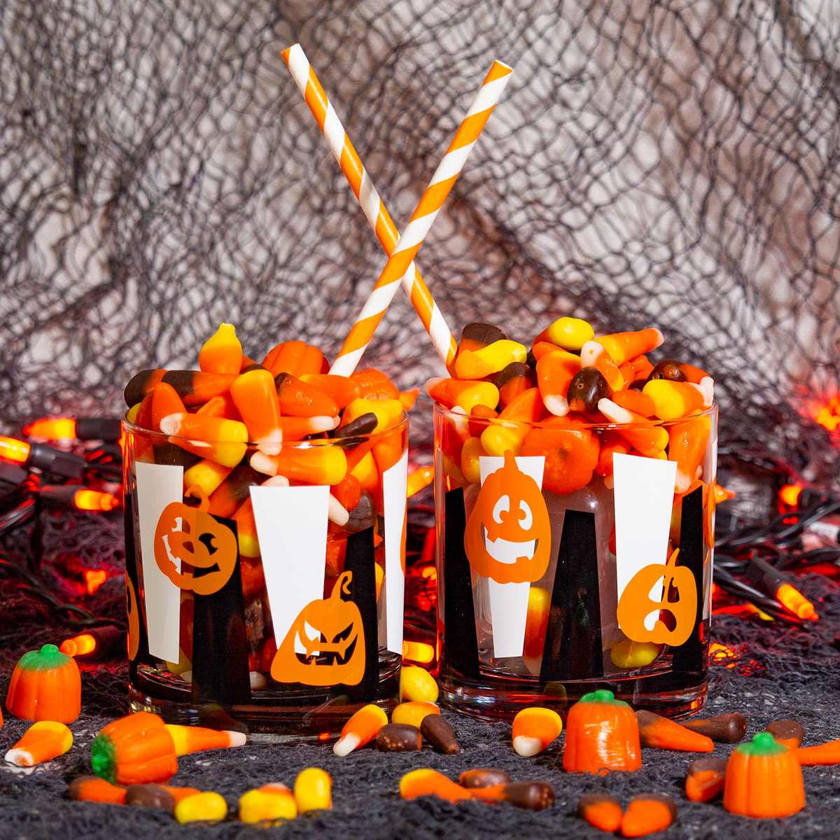 Two Halloween-themed Rocks drinkware filled with candy corn and chocolate candies, handprinted with orange jack-o'-lanterns over black and white vertical googies, each topped with an orange-and-white striped paper straw. The cups are set on a dark surface with scattered candy, small pumpkin decorations, and a spooky web-like backdrop.