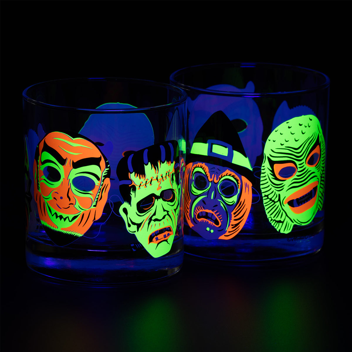 Two lined up Neon Nightmares Rocks glasses with screen printed retro monster masks glowing fluorescent under a blacklight on a black background. 