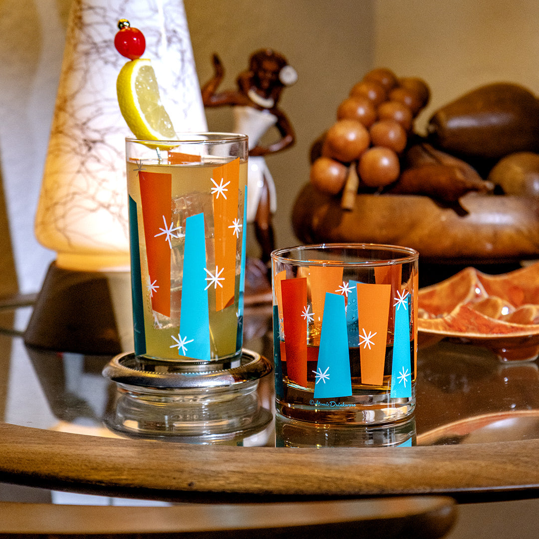Two colorful cocktails in Palm Springs glass tumblers on a wooden table with decorative elements in the background.